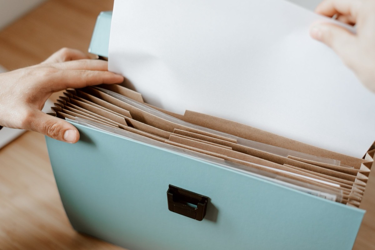 Hands lifting a blank sheet out of an accordion file folder with brown dividers, the kind of sorting that comes before papers from Pakistan are sent for attestation