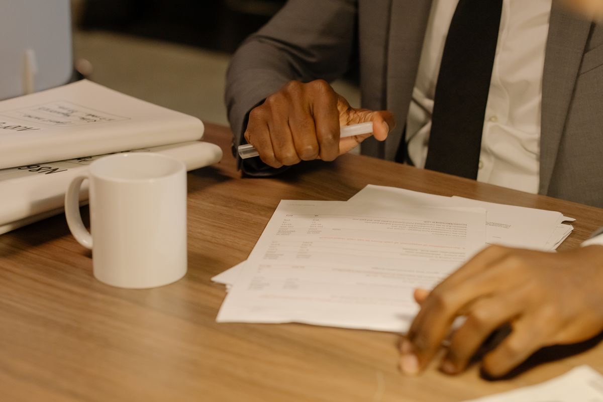 A man in a grey suit and dark tie at a wooden desk going through a small stack of printed pages, with a white mug and bound files beside him, like a company officer gathering the parent company's papers
