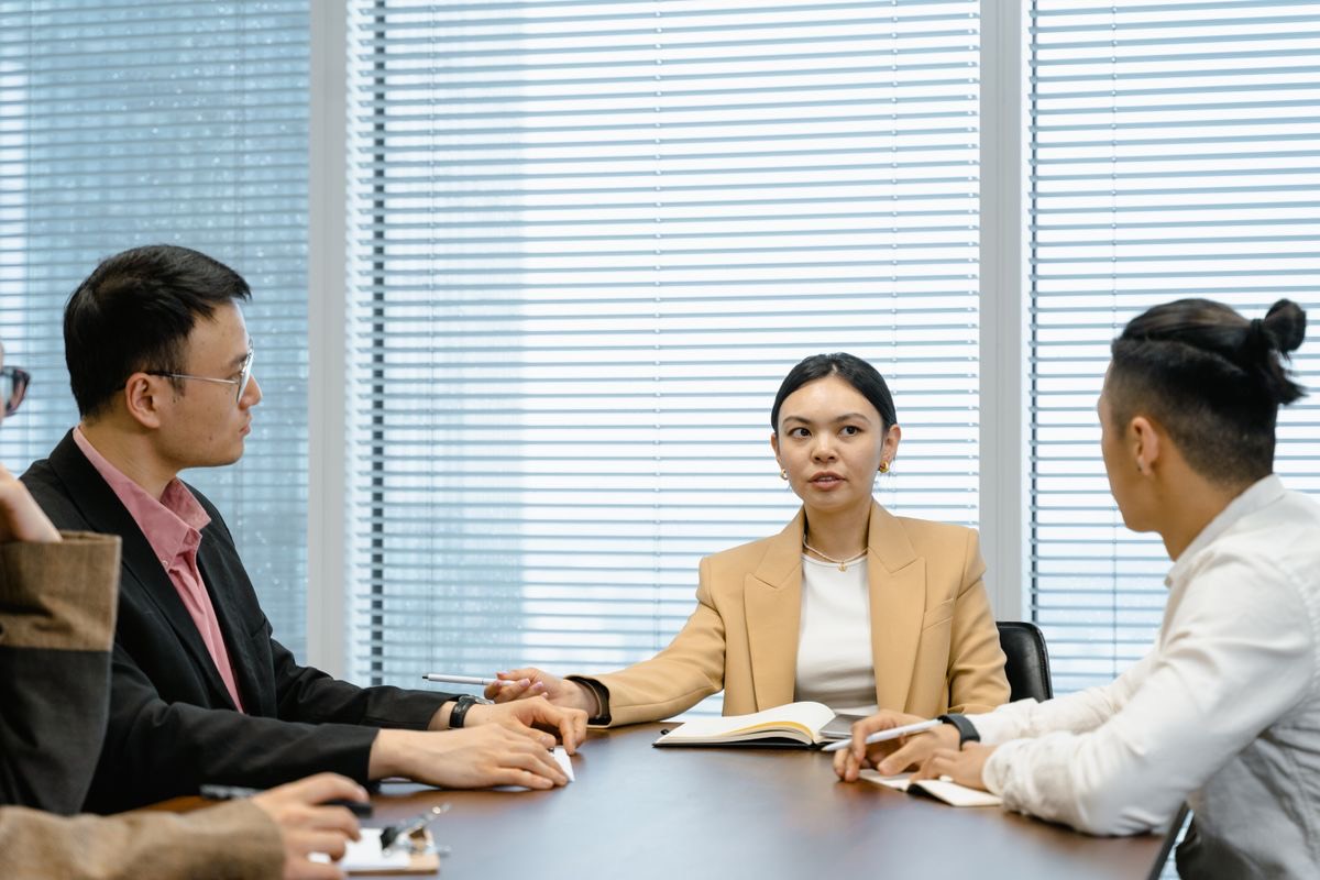 Four colleagues around a conference table in an office with window blinds, a woman in a beige blazer speaking while the others listen with notebooks open, like a parent company's managers choosing how to set up in Dubai