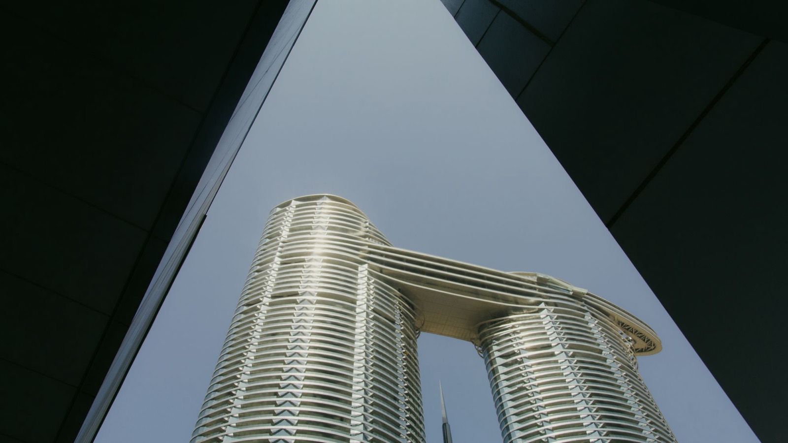 Two towers in Dubai joined near the top by a sky bridge, seen from street level between two dark buildings, like a parent company and the Dubai entity that stays linked to it