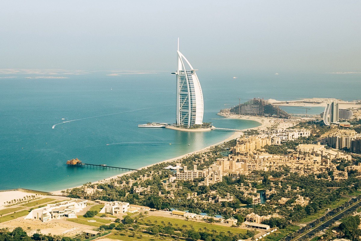 Aerial view of the Dubai coastline at Jumeirah, with the sail shaped Burj Al Arab hotel standing offshore, a long beach and low rise resort buildings among palm trees, the Dubai side of the comparison table in this section