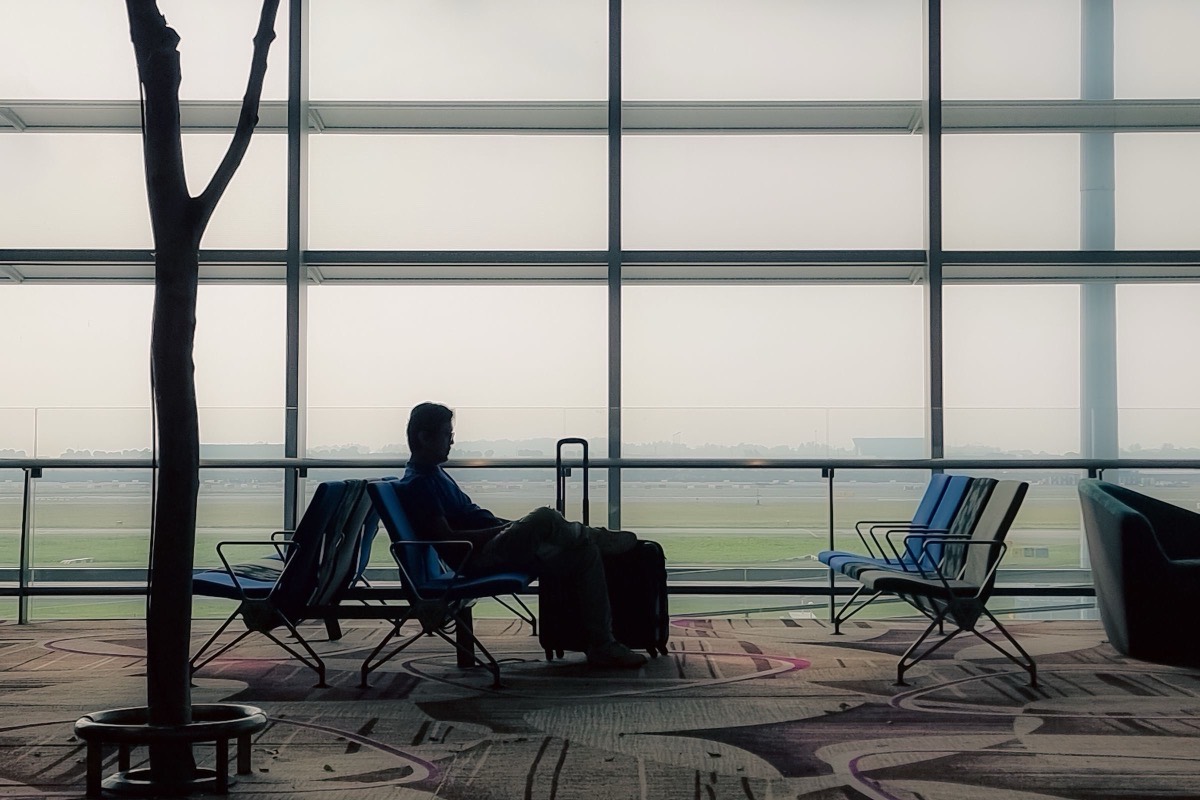 A traveller in silhouette sits with a suitcase in an airport lounge beside tall windows looking out over the runway, a picture of a founder weighing a move abroad with a new company