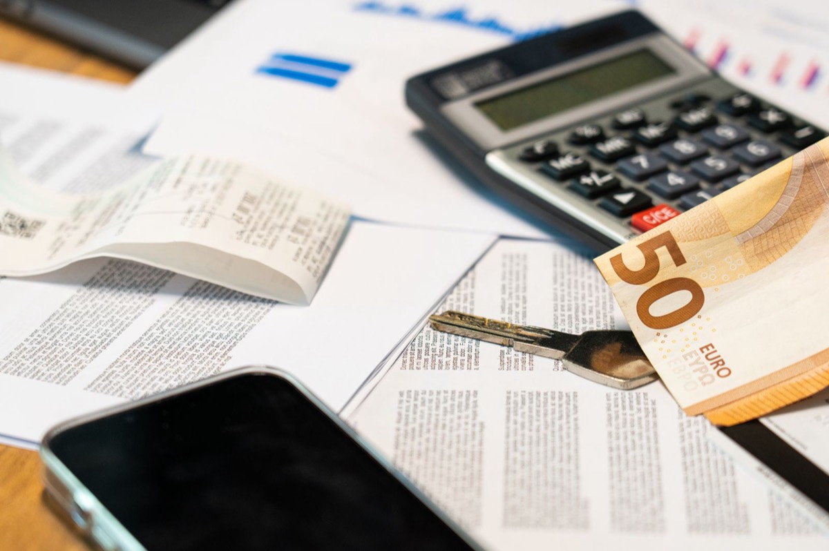 A 50 euro banknote, a calculator, a till receipt, a key and a phone lying on printed papers on a desk, like the household paperwork you sort out before leaving Spain
