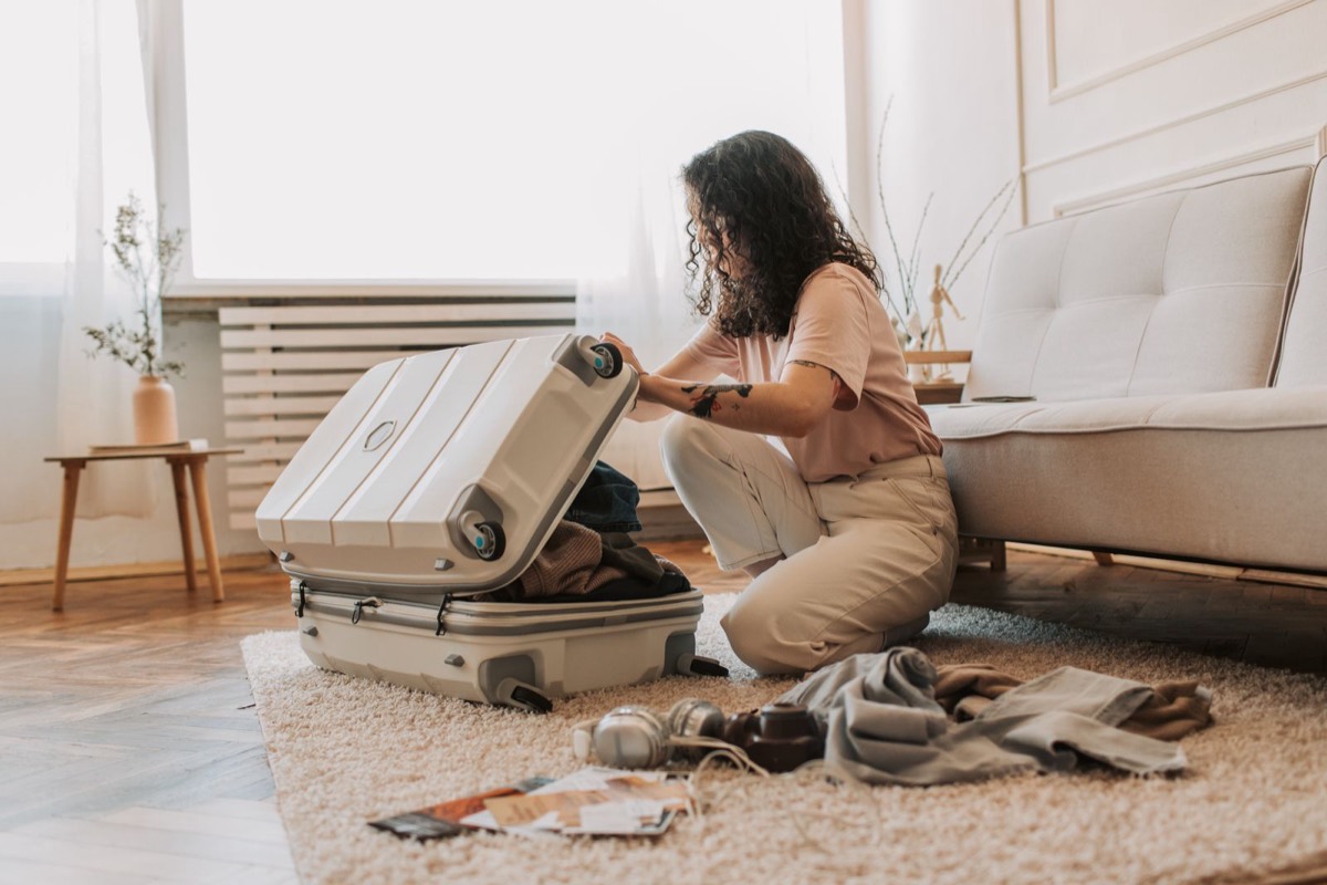 A woman kneeling on a rug at home and opening a white suitcase half full of clothes, with headphones, a camera and papers on the floor, getting ready for a move abroad