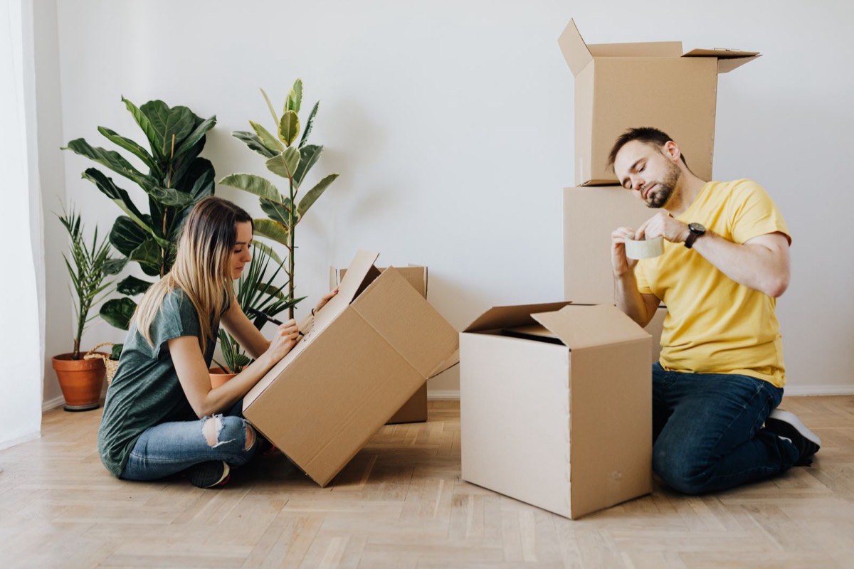 A man and a woman sitting on a wooden floor packing and labelling cardboard boxes in an empty room, getting ready to move abroad